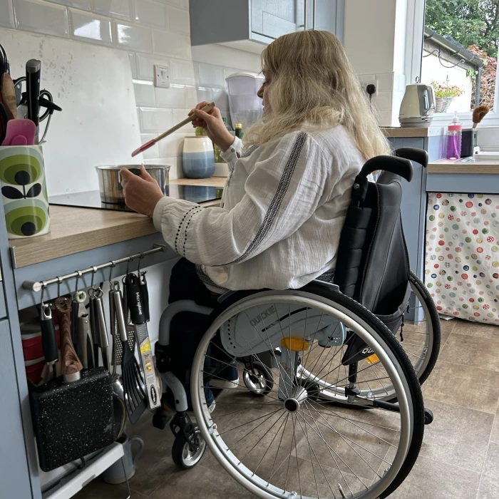 a woman sitting in a wheelchair is stirring a saucepan the hob is height adjustable with space below to allow her to sit close the kitchen units are pale blue with wooden worktops she has long blonde hair and is wearing a white top