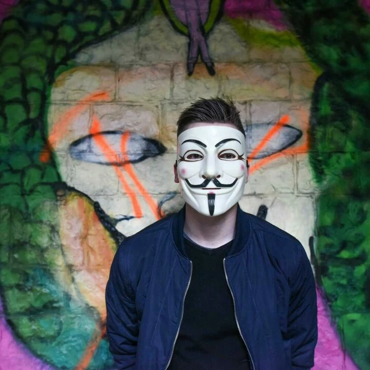 A person in a Guy Fawkes mask stands before colorful graffiti art, creating an air of mystery.