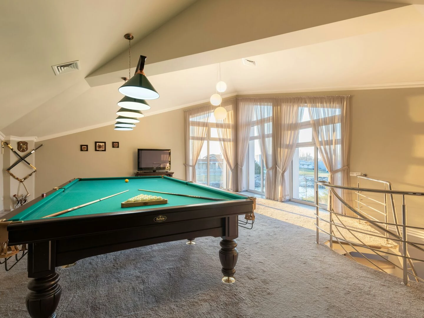 wooden pool table with cues placed on carpet in spacious room with windows and staircase with metal railing in contemporary building